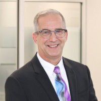 A man with short gray hair and glasses is smiling. He is wearing a black suit, white shirt, and a colorful tie. The background features light-colored walls and frosted glass panels.