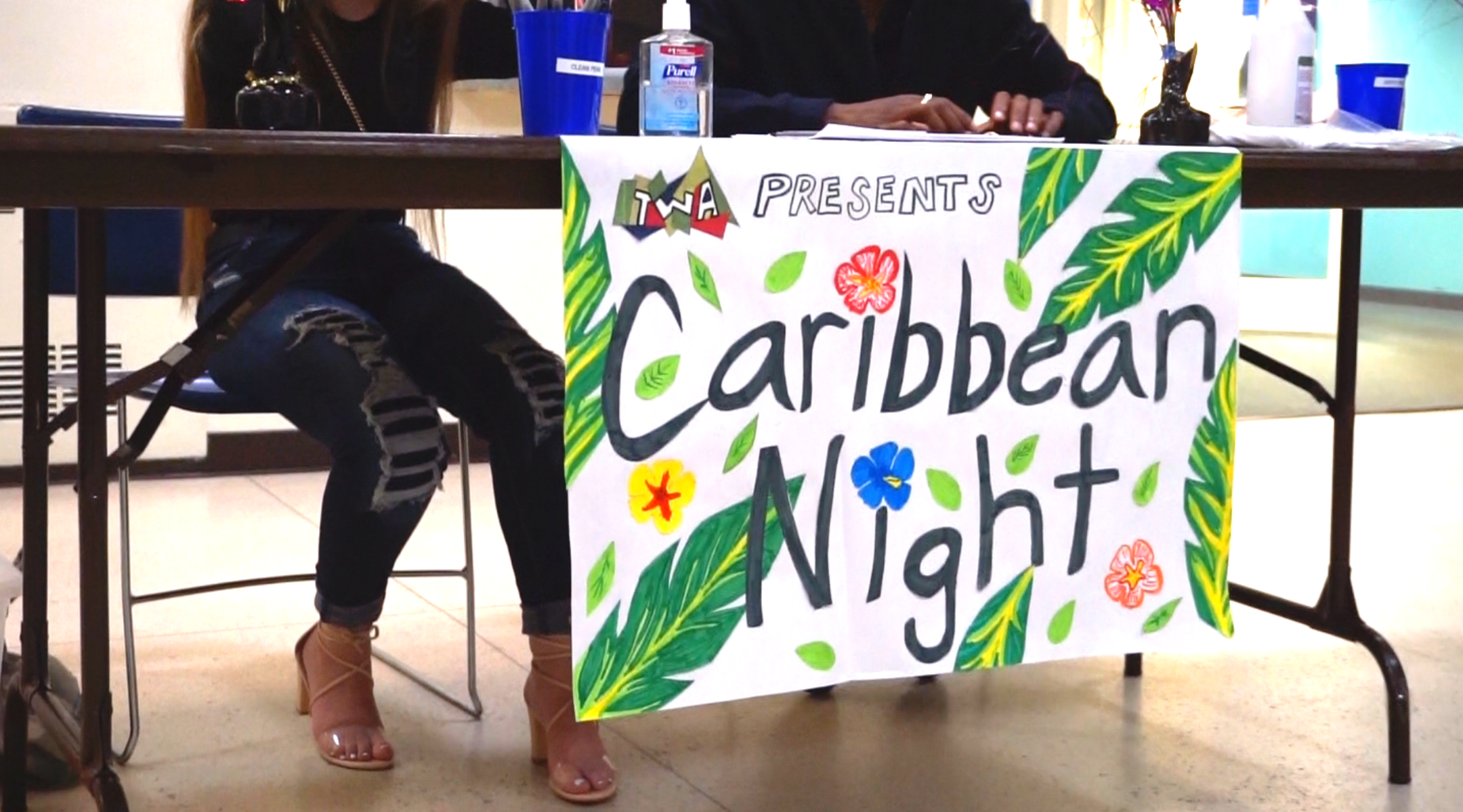 A poster sign for Caribbean night at Worcester State taped to a table with students sitting behind it