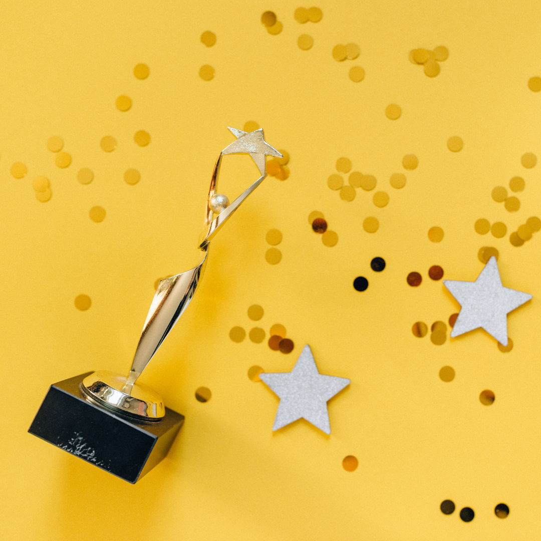 A gold star-shaped trophy on a black base sits on a yellow surface with scattered gold confetti and two white star cutouts.