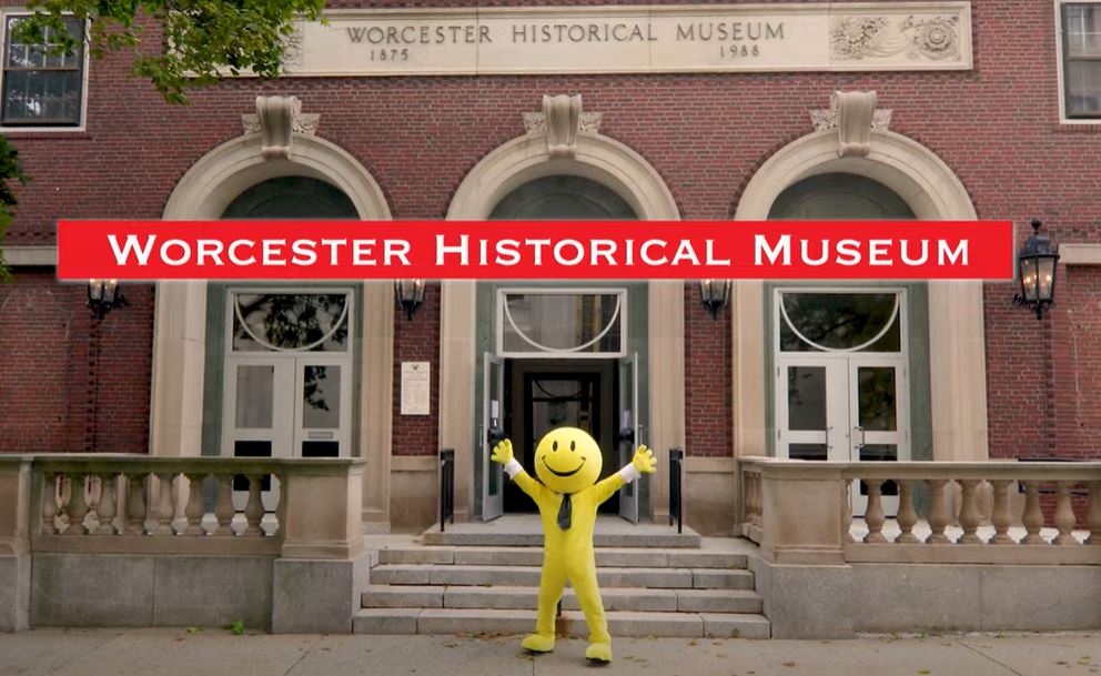 A smiley-face mascot standing with it's arms spread in front of the Worcester Historical Museum