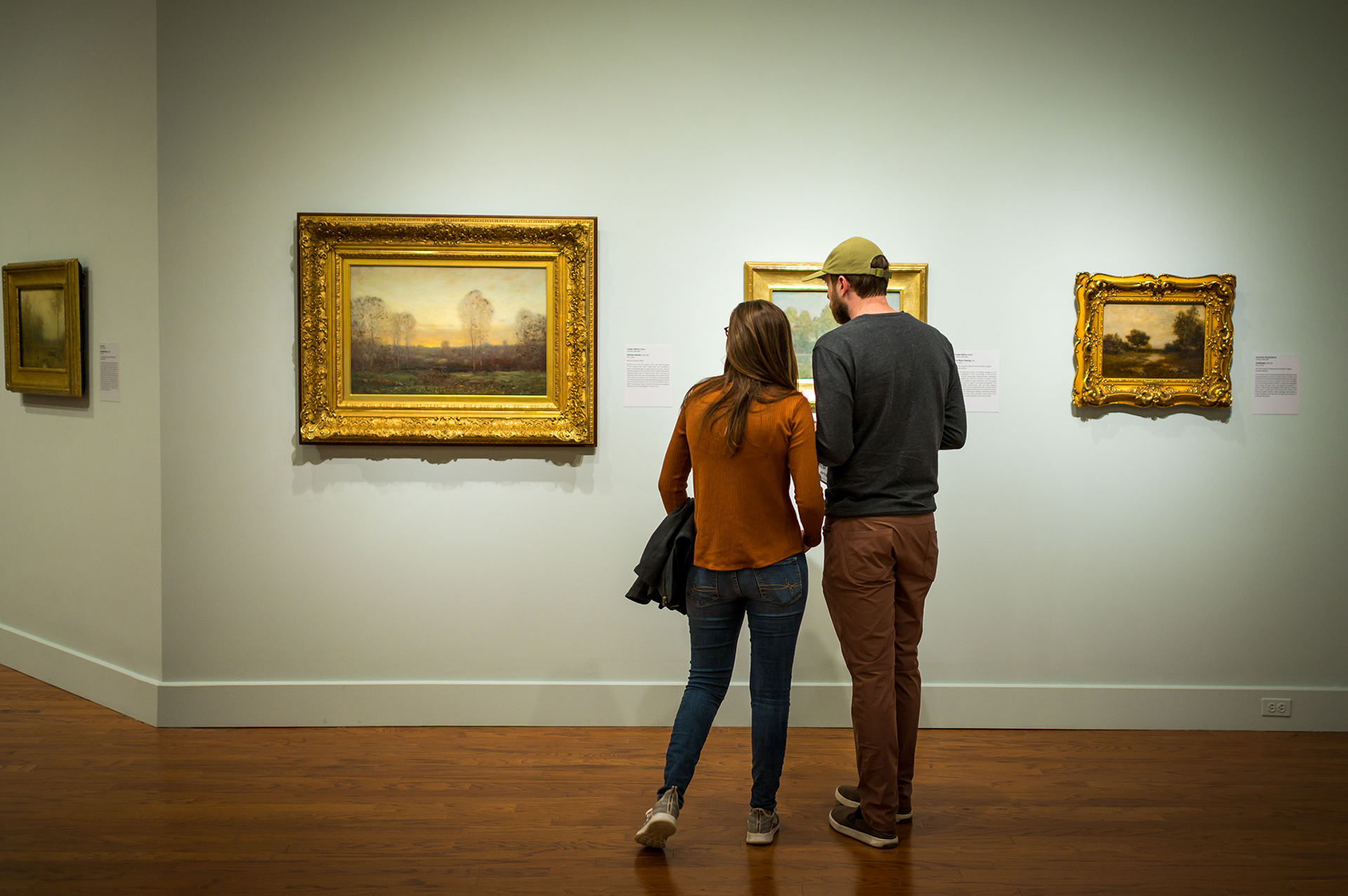 two individuals with their backs to the camera looking at landscape paintings in golden frames hanging on the wall at the Worcester Art Museum