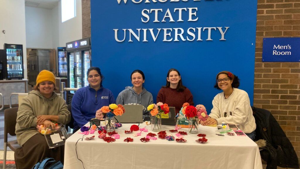 Five people sit at a table decorated with colorful artificial flowers, smiling at the camera. Behind them is a blue sign reading Worcester State University. A vending machine and mens restroom sign are visible.