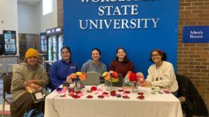 Five people sit at a table decorated with colorful artificial flowers, smiling at the camera. Behind them is a blue sign reading Worcester State University. A vending machine and mens restroom sign are visible.