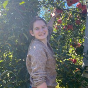 A woman smiles at the camera while reaching up to pick a red apple from a tree in a sunny orchard, surrounded by green leaves and ripe apples.