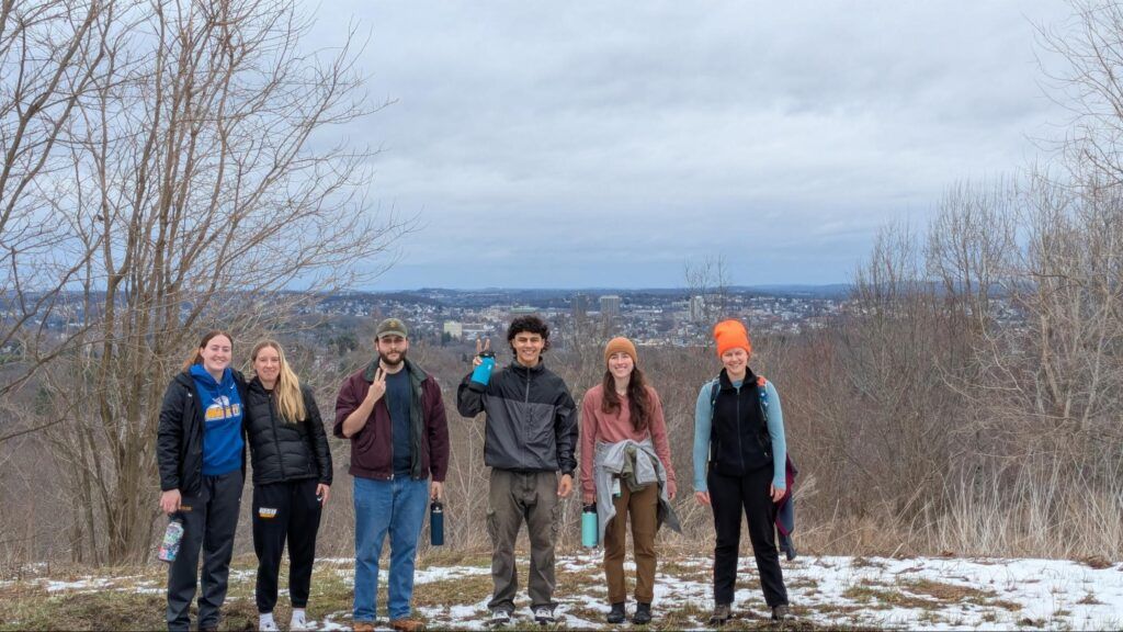 Six people in winter clothing stand on a snowy hilltop with bare trees, overlooking a distant city under a cloudy sky.