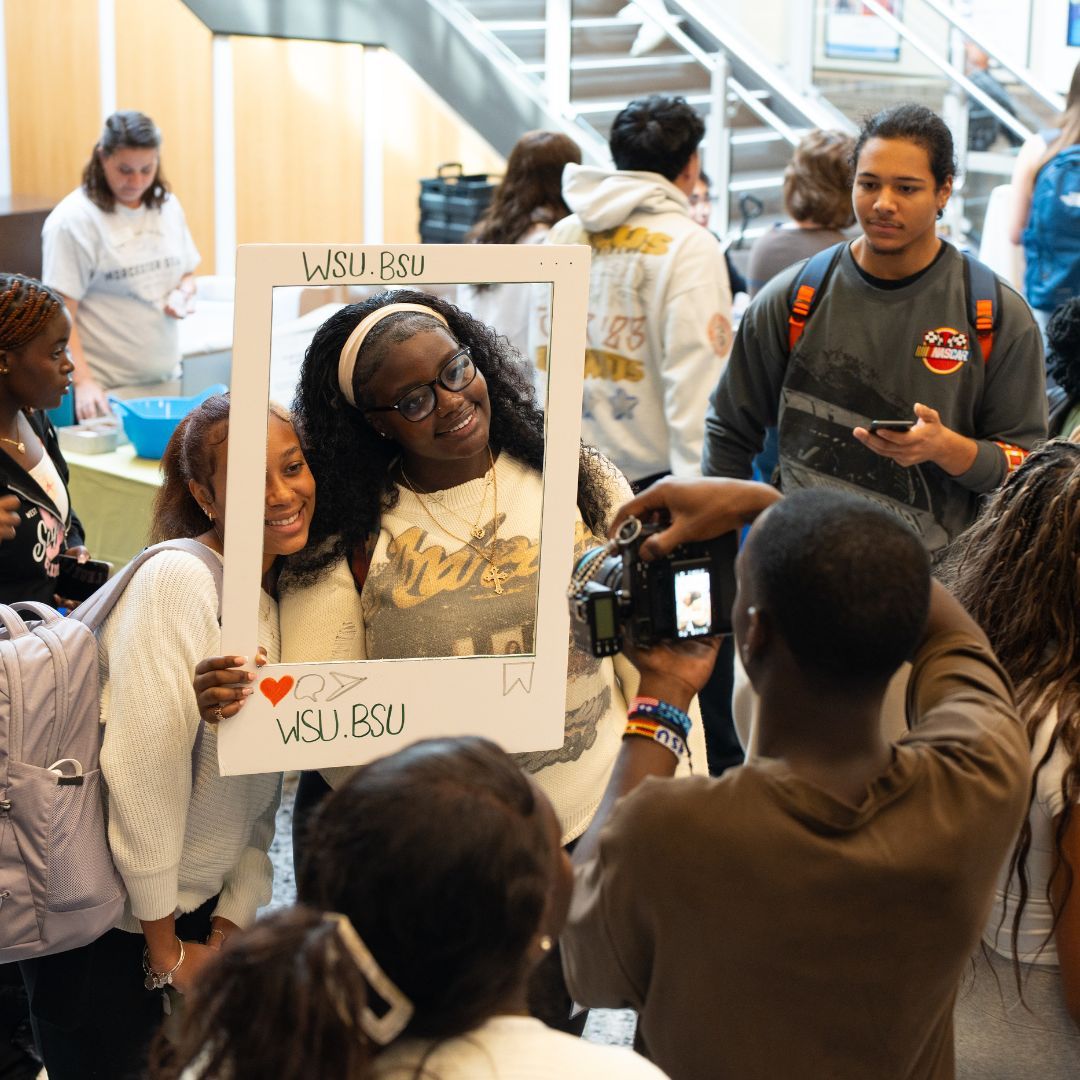 A group of students gathers indoors; two pose smiling with a WSU.BSU frame while another takes their photo. Others mingle in the background.