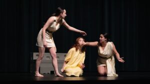 Three women in light-colored costumes perform on stage; one is kneeling, one is seated being held by the chin, and the third stands with an arm outstretched toward them.