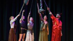 Five people in fantasy costumes stand in a row on stage, holding up swords and weapons together, with a dark curtain backdrop.