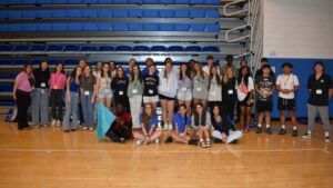 A group of young people pose for a photo in a gymnasium, standing and kneeling in front of blue bleachers.