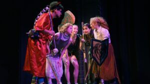 Five people in costume stand close together on stage, holding props like swords and shields, appearing to be in an intense or dramatic conversation during a performance.