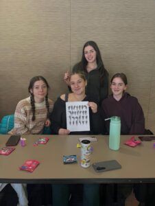Four young women sit at a table with snacks and drinks; one holds up a paper with black-and-white illustrations.