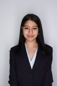 A young woman with straight dark hair, wearing a navy blazer and light blue blouse, stands against a plain light gray background.