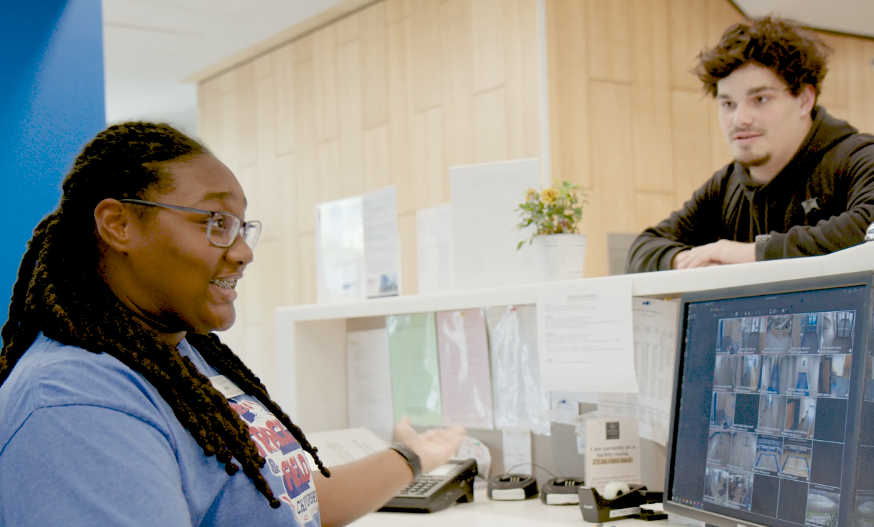 A woman at a reception desk speaks to a man leaning on the counter; the desk has a computer monitor showing security camera feeds.
