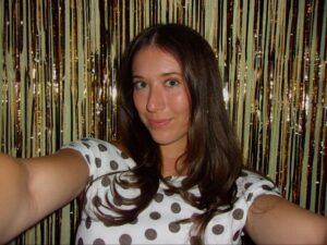 A woman with long brown hair wearing a white shirt with black polka dots takes a selfie in front of a gold tinsel backdrop.