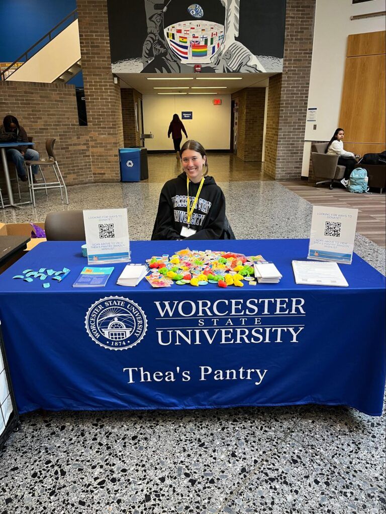 A person sits behind a table covered with items and informational materials for Thea's Pantry at Worcester State University, with a blue tablecloth displaying the pantry’s name.