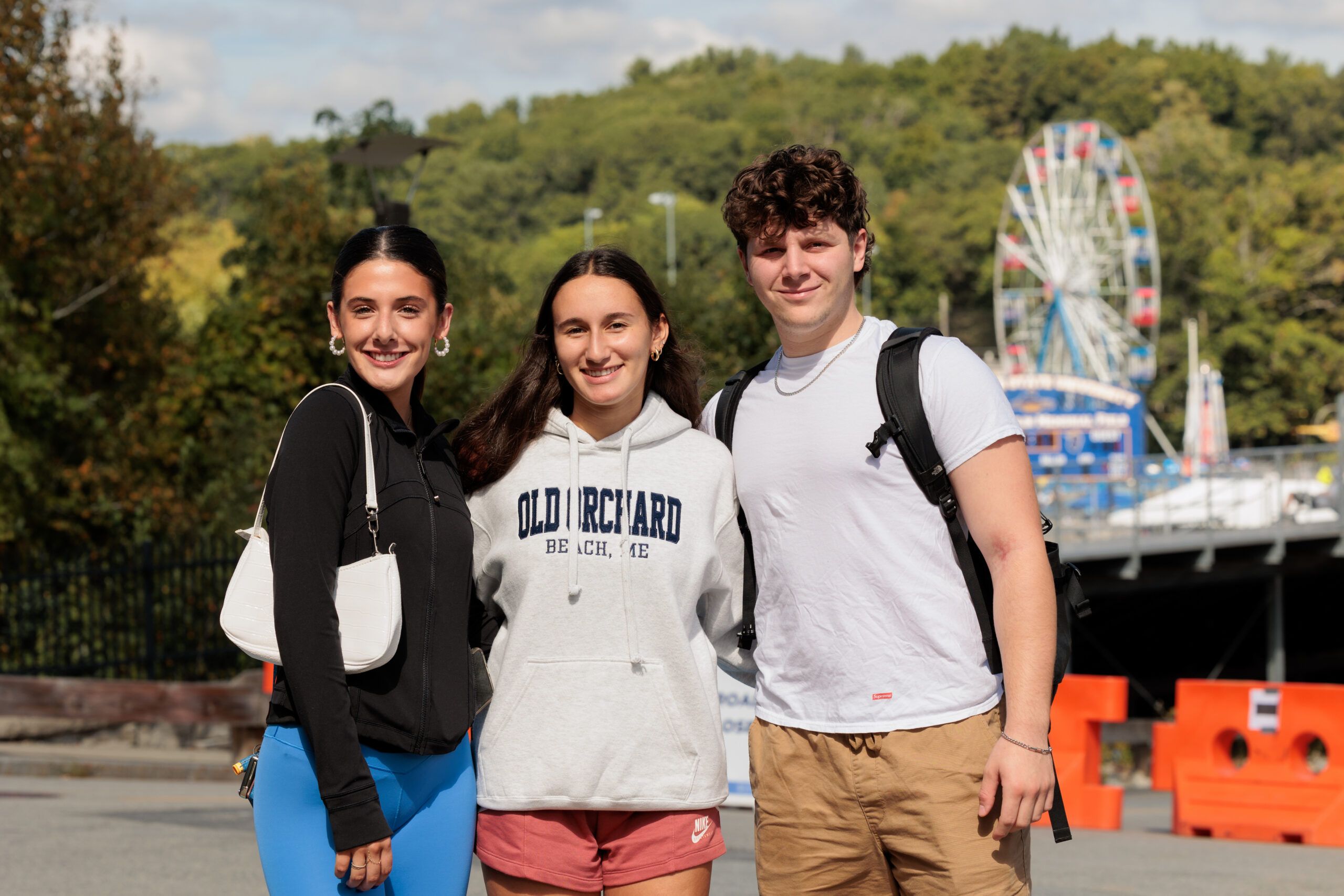 Three young people stand outdoors smiling, with trees and a Ferris wheel in the background on a sunny day.