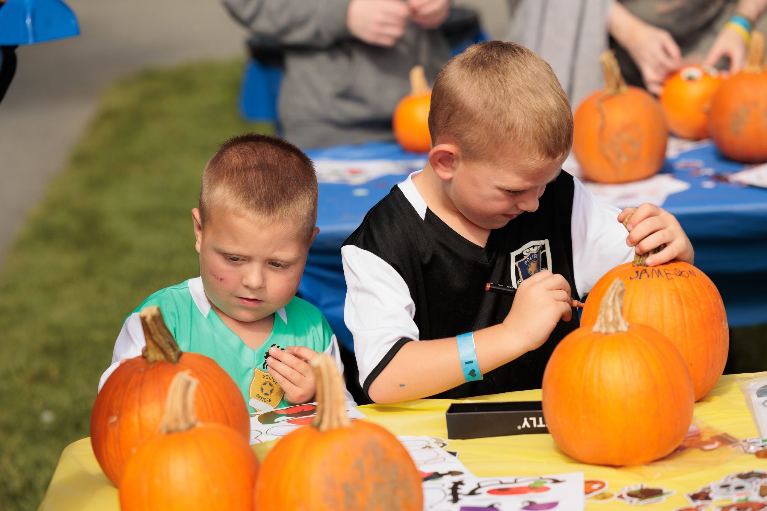 Two young boys sit at a table outdoors, decorating small pumpkins with markers. Several pumpkins and art supplies are on the table around them.
