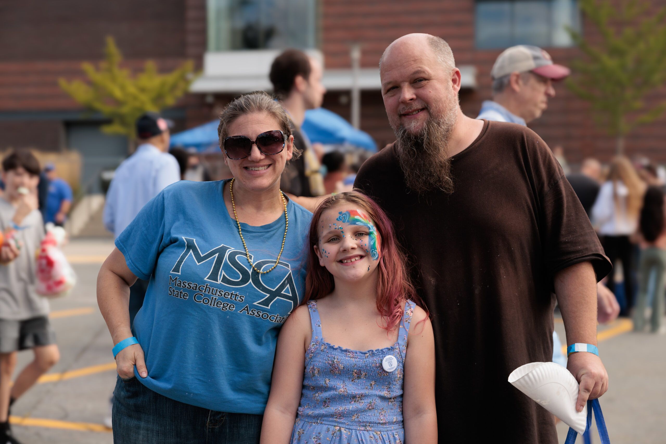A woman, a man, and a girl with face paint smile outdoors at an event. The girl wears a blue dress; others are visible in the background.