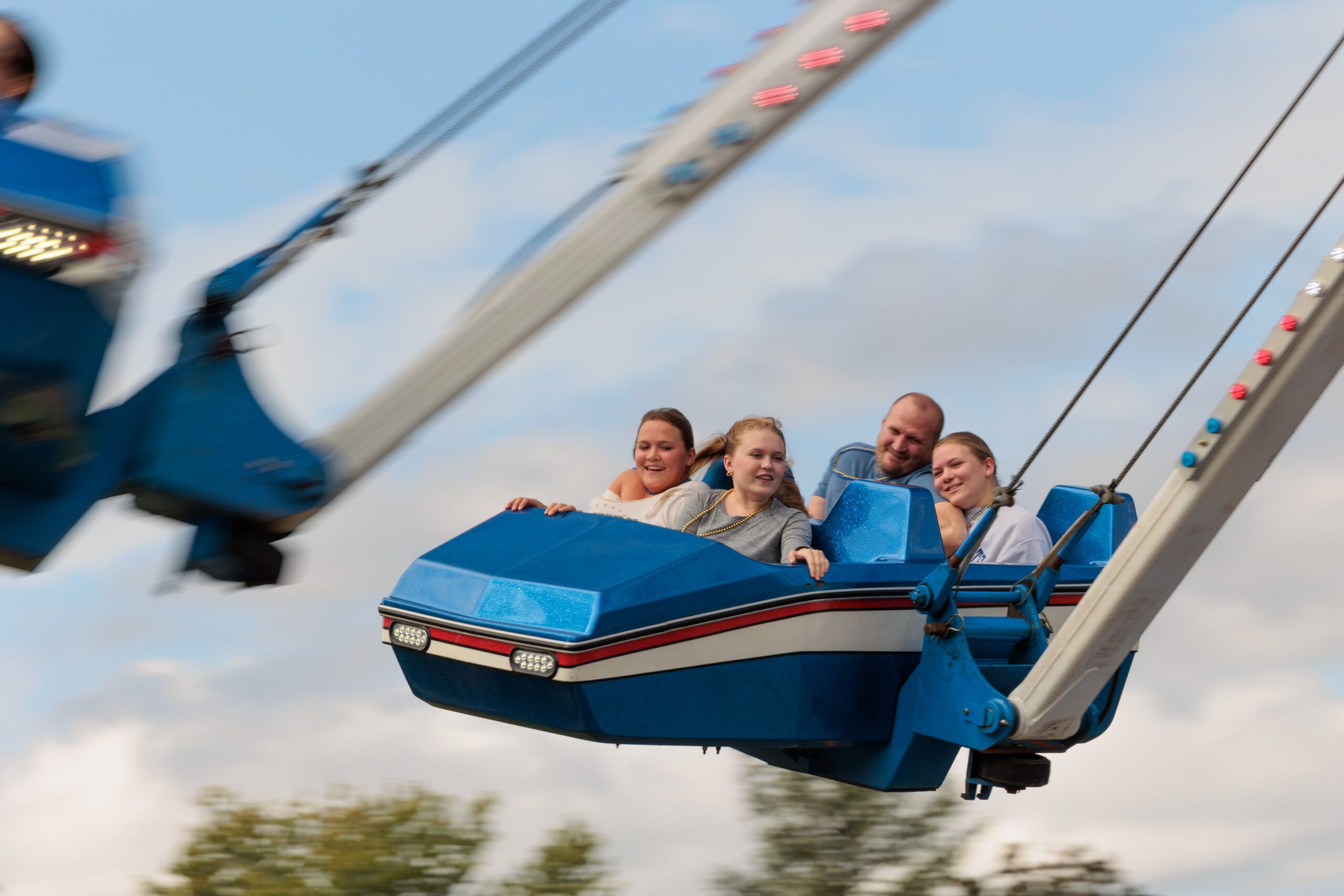 Four people ride a blue amusement park swing ride, smiling and holding onto the safety bars as the ride moves through the air against a partly cloudy sky.