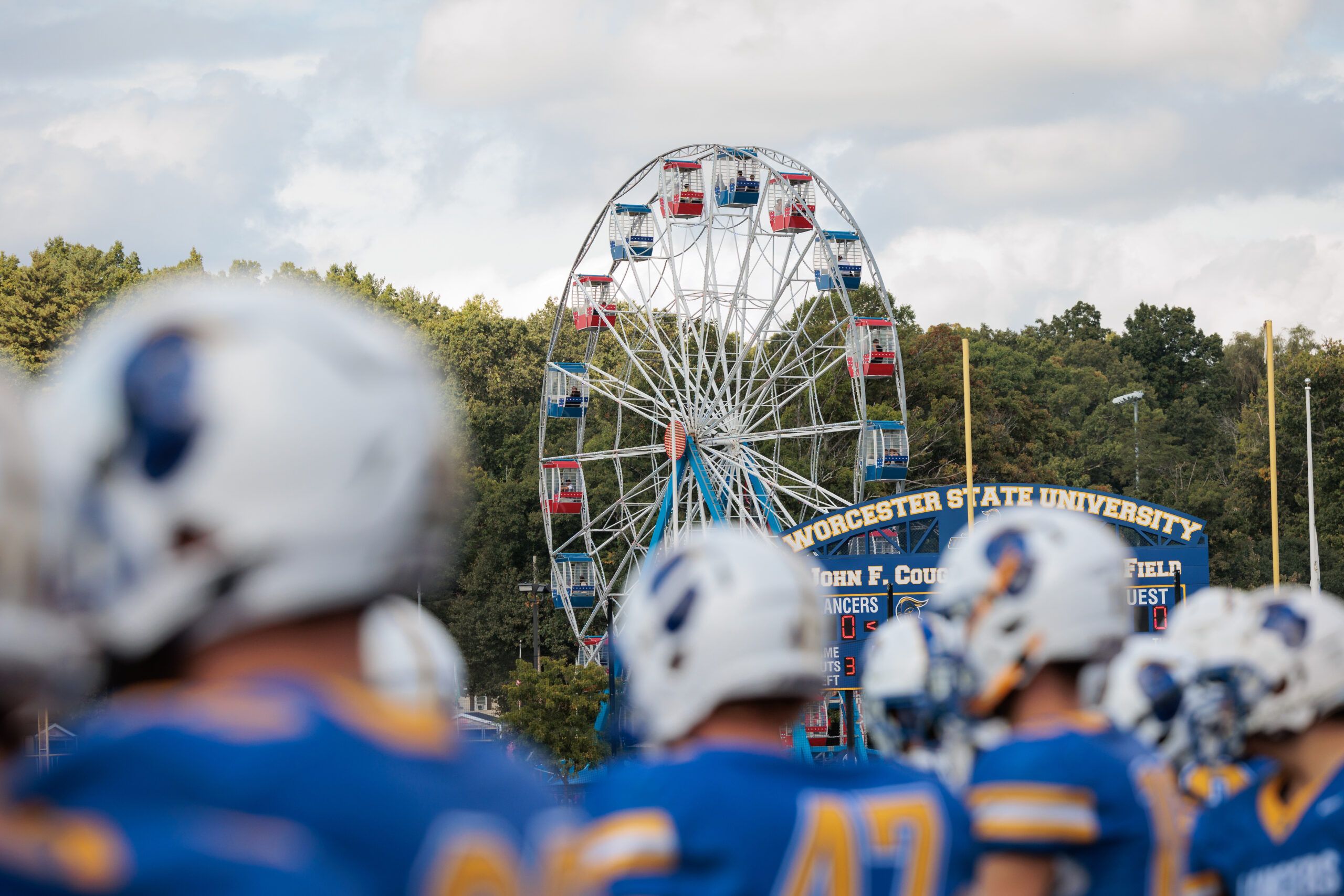 A Ferris wheel rises behind a scoreboard and blurred football players in blue uniforms at Worcester State University.
