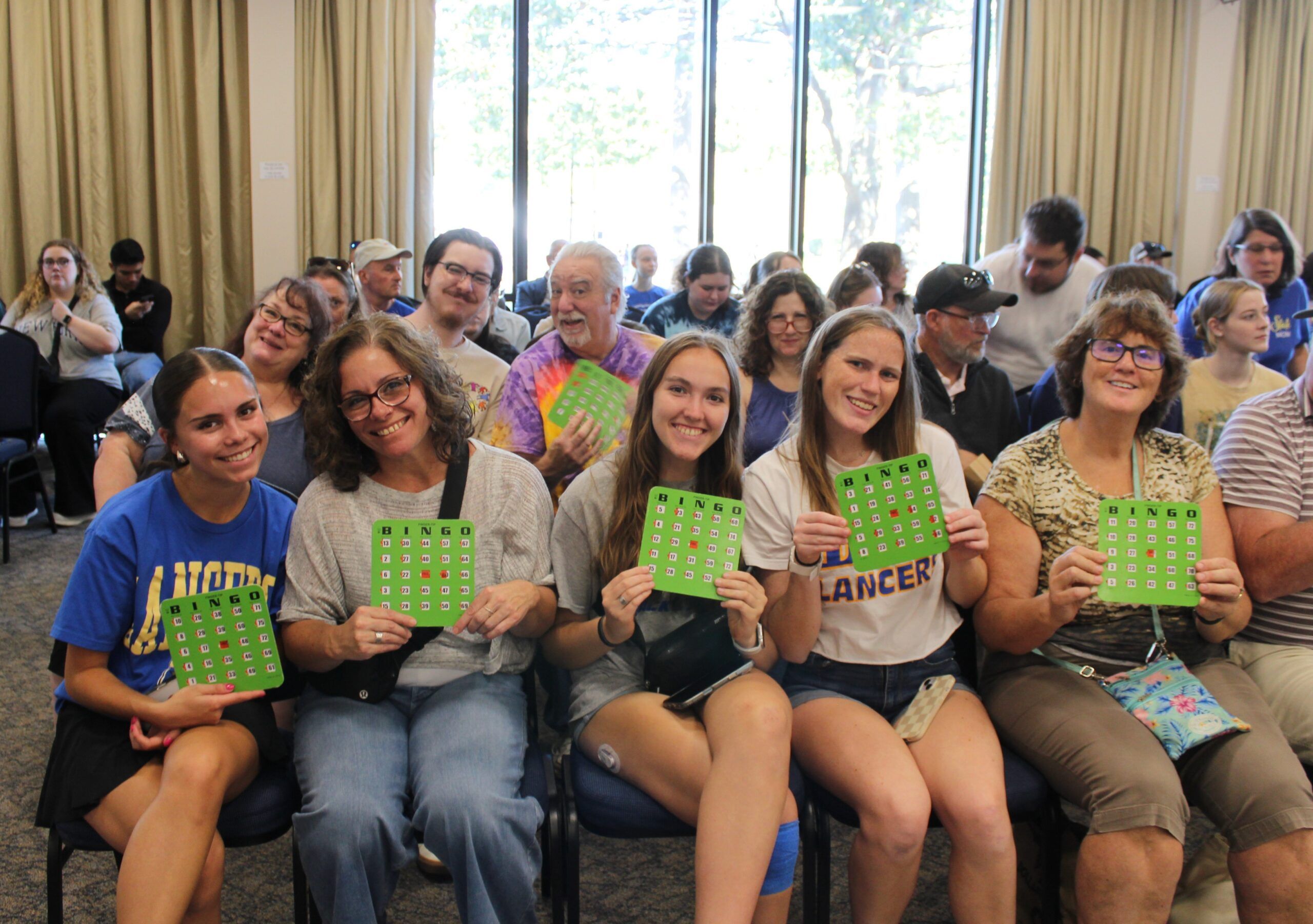 A group of people sit in rows indoors, smiling and holding green bingo cards, with large windows and trees visible in the background.