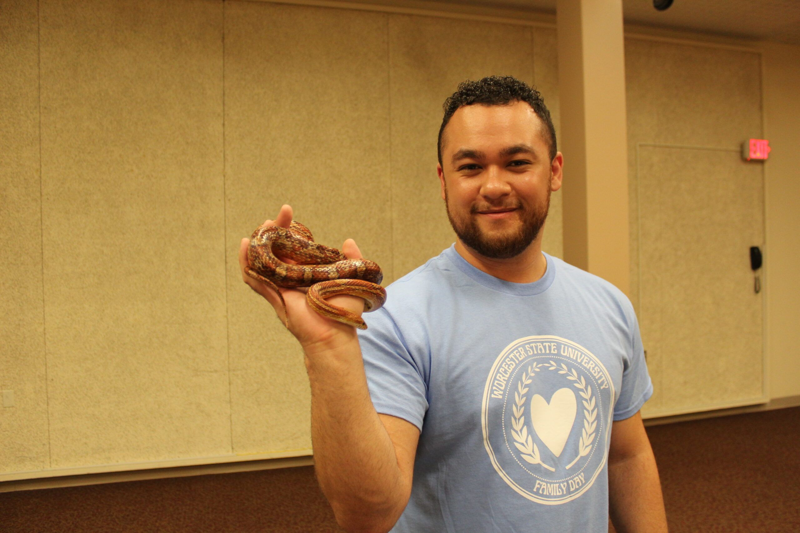 A man in a blue university t-shirt holds a small, coiled snake in his left hand while standing indoors.