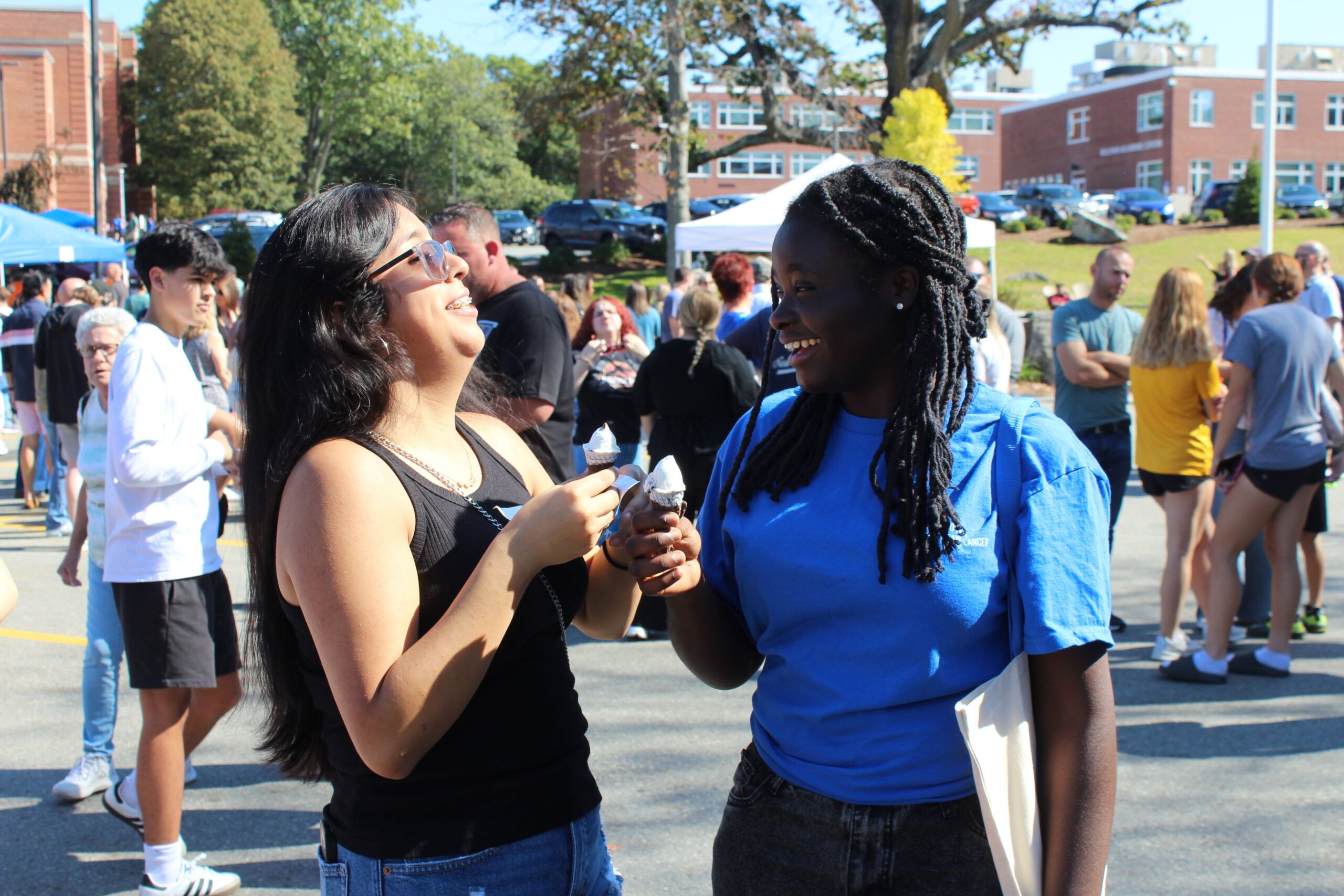 Two people stand outside at a crowded event, smiling and holding ice cream cones, with buildings and parked cars visible in the background.