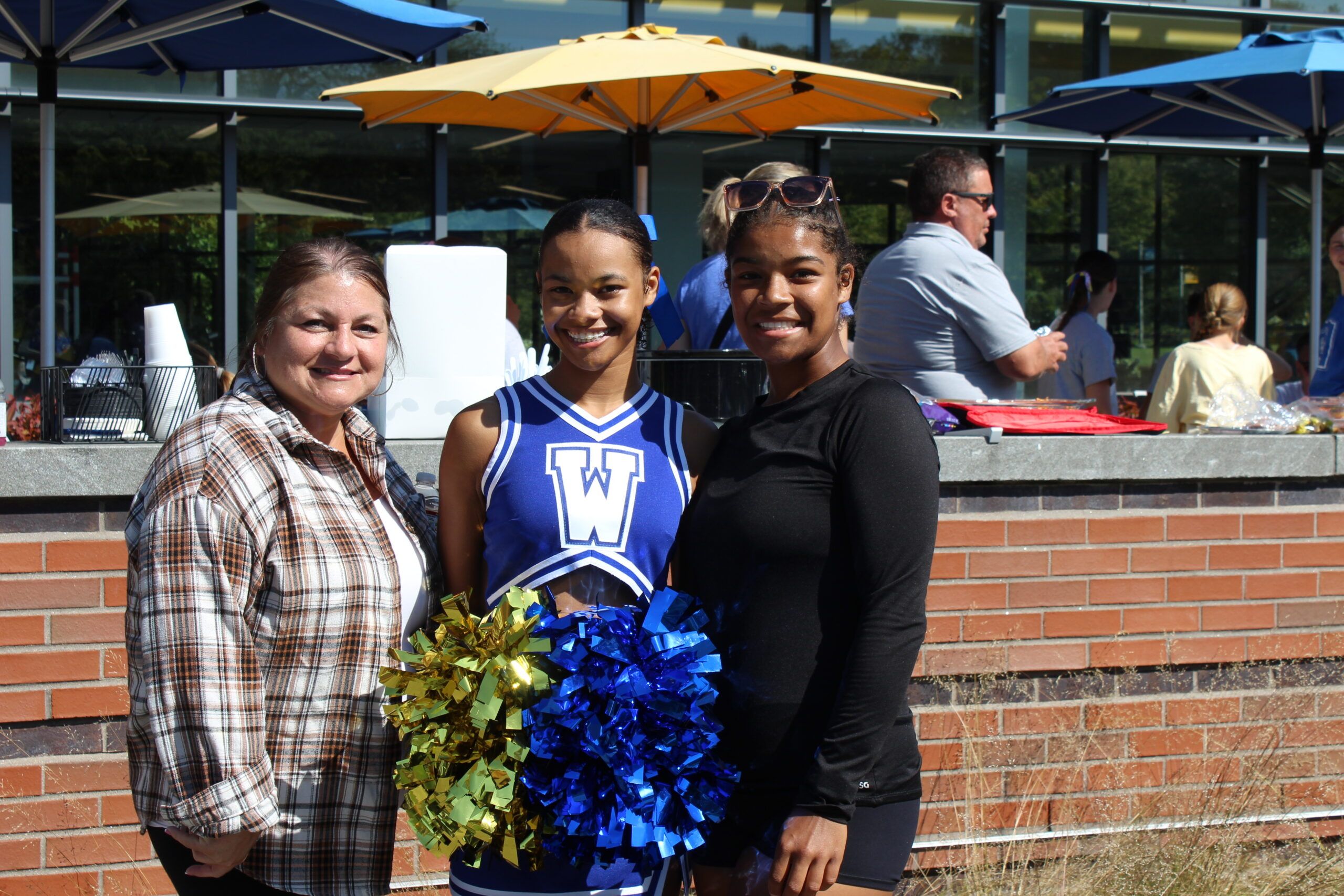 Three women pose together outdoors; one is in a blue cheerleader uniform holding yellow and blue pom-poms. Several people are visible in the background near a brick wall and umbrellas.