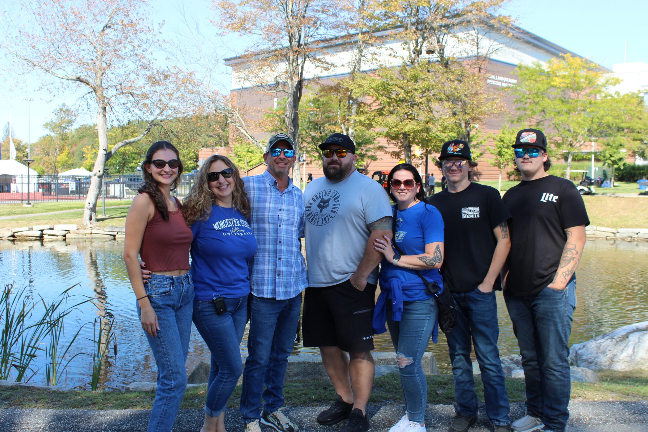 Seven adults stand side by side, smiling outside near a pond with trees and a large brick building in the background.