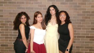 Four women stand in front of a brick wall, posing for a group photo. Three wear skirts or dresses, one wears glasses and a smart watch.