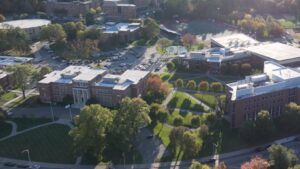 Aerial view of a college campus with multiple buildings, parking lots, trees, and a sports field on a sunny day.