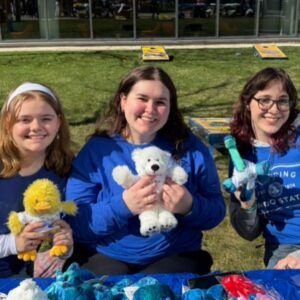 Three people sit outside at a table holding stuffed animals, with a grassy area and games in the background.