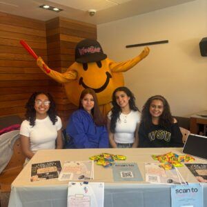 Four women sit at a table with flyers and candy, promoting an event. Behind them stands a person in a yellow mascot costume wearing a cap.