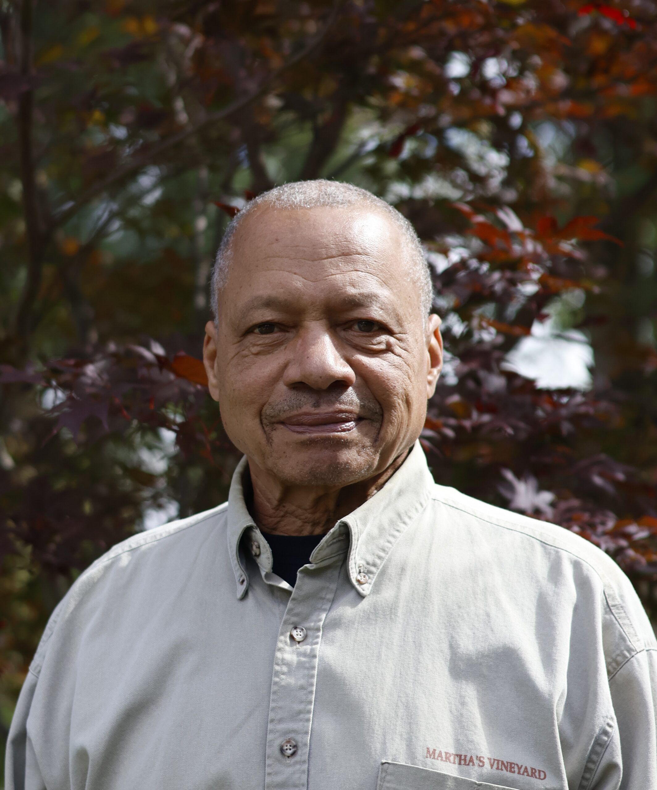 An older man with short gray hair stands outdoors in front of leafy trees, wearing a light-colored button-up shirt that reads “Martha’s Vineyard.”.