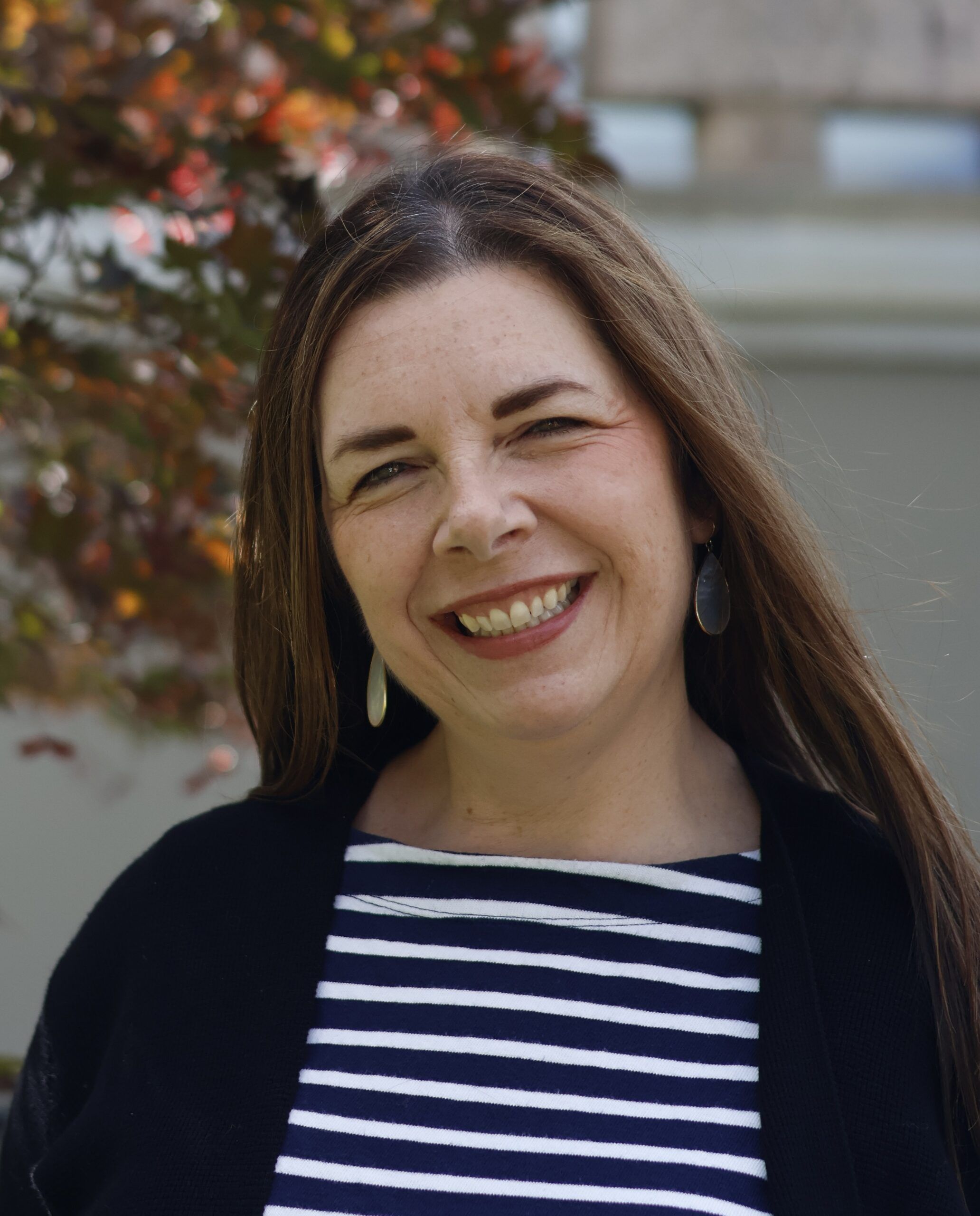 Woman with long brown hair, wearing a black sweater and striped shirt, smiles outdoors in front of a leafy background.