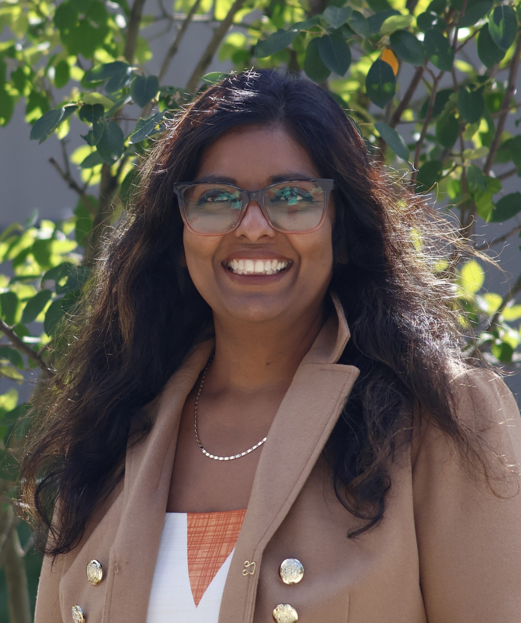 A woman with long dark hair and glasses smiles at the camera outdoors, wearing a tan coat over a white and orange top, with green foliage in the background.
