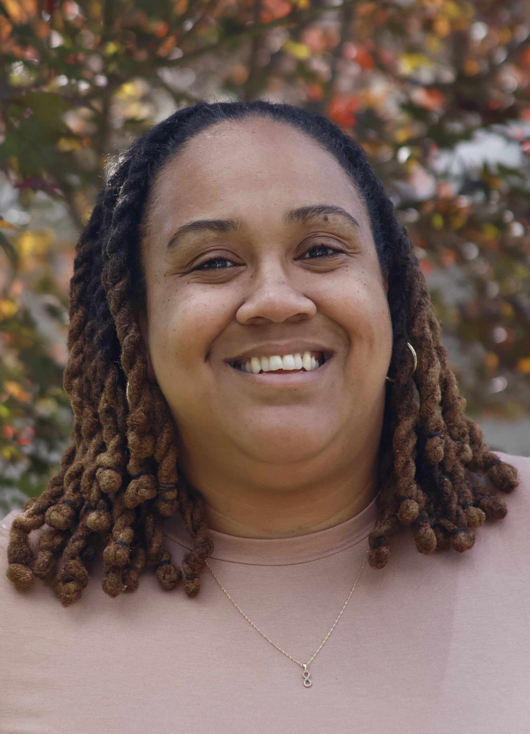 A person with medium-length locs and a light brown shirt smiles outdoors, with blurred foliage in the background.