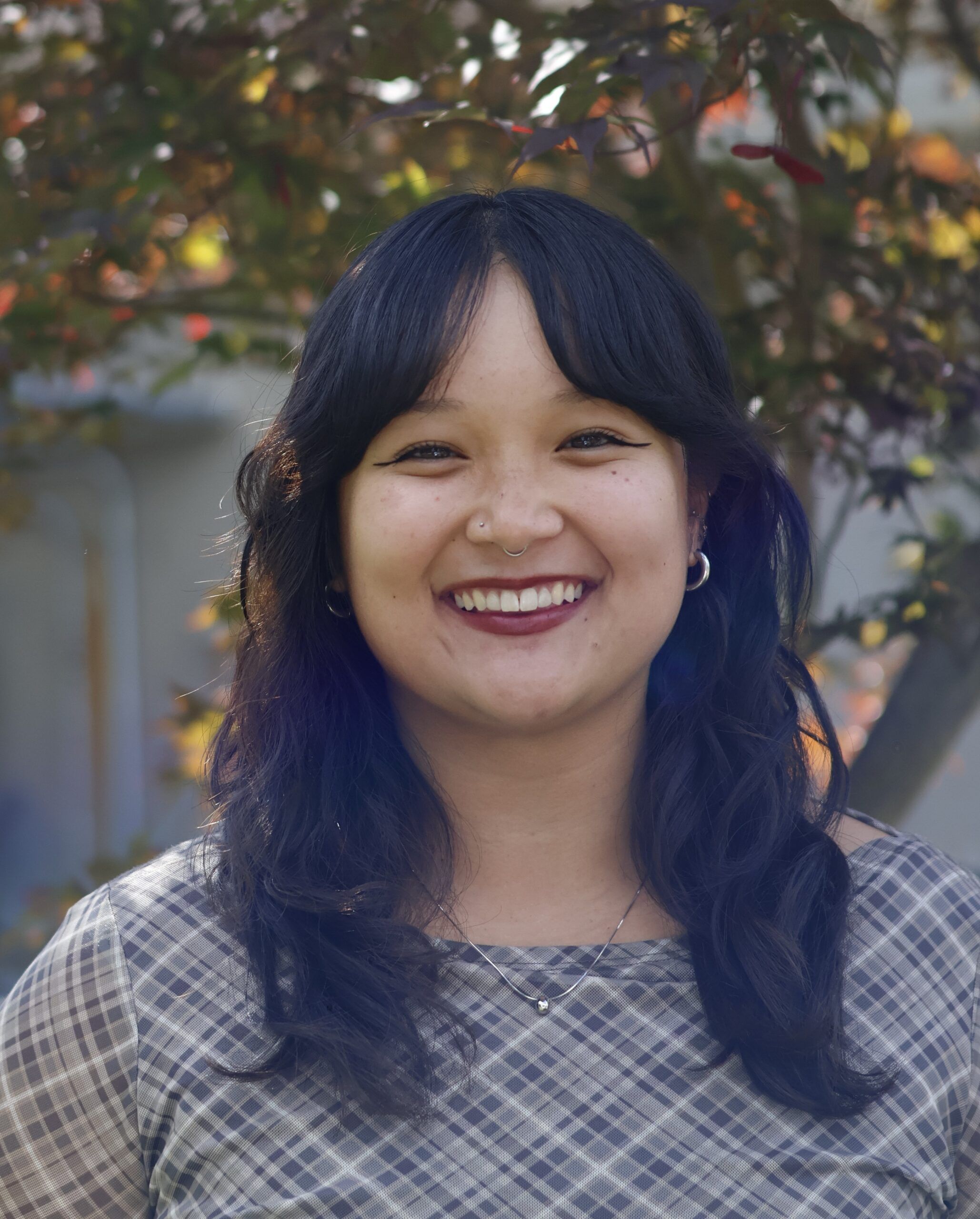 A woman with wavy dark hair and a plaid shirt smiles outdoors, with sunlight and foliage in the background.