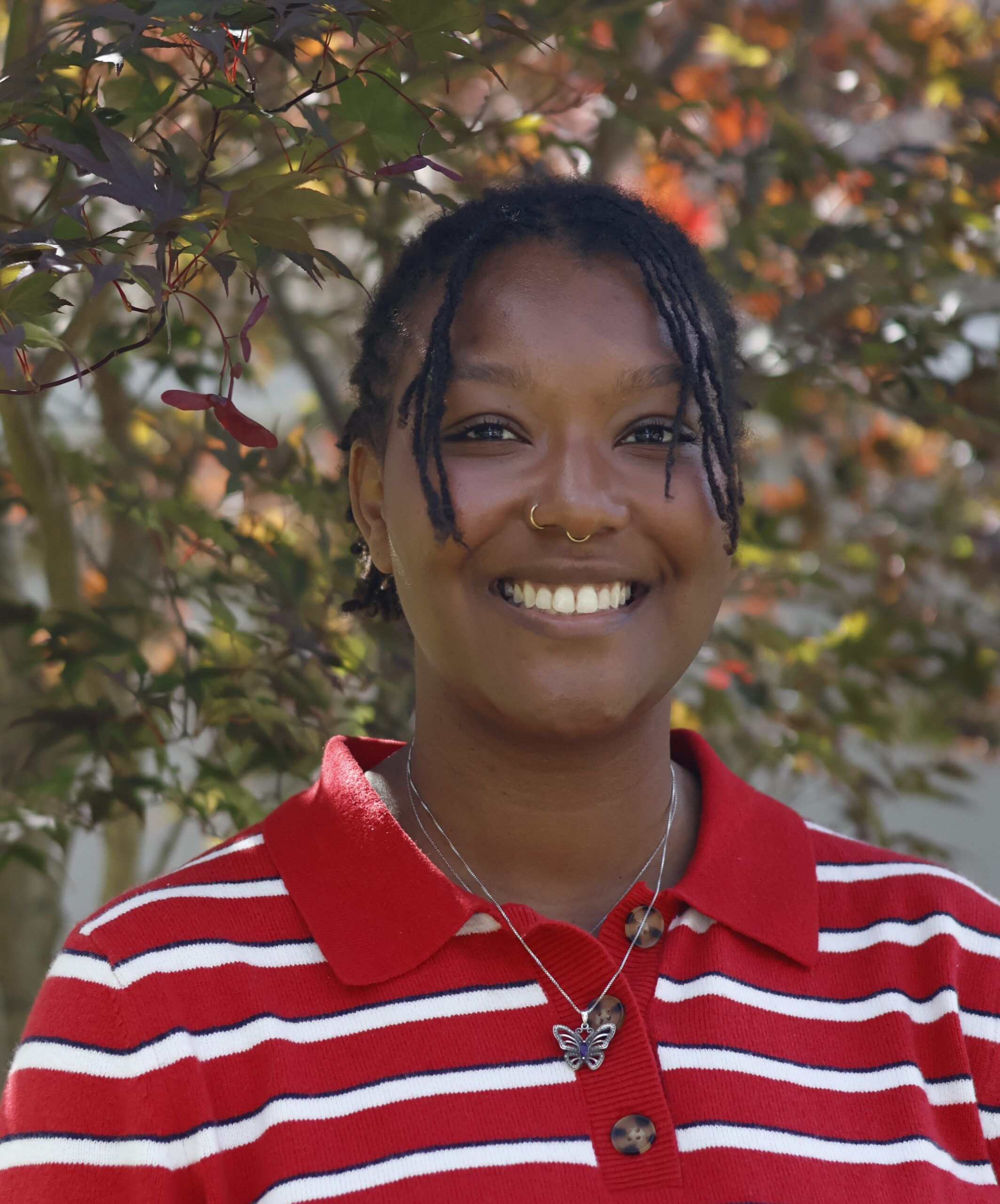 A person with short braids, wearing a red and white striped shirt and a butterfly necklace, smiles in front of leafy branches outdoors.