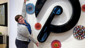 A woman stands next to a large, black, three-dimensional Pinterest logo on a white wall decorated with circular paper ornaments.