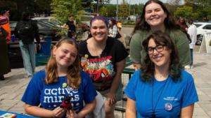 Four young women smiling at an outdoor event, with a table in front of them and people in the background.