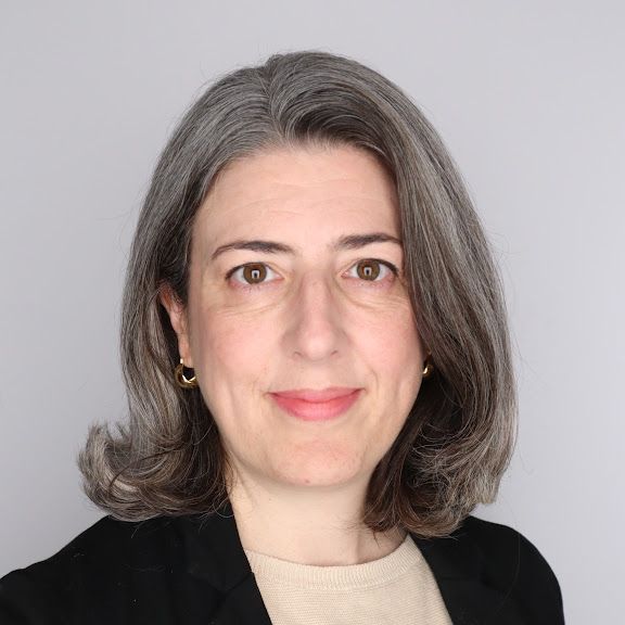 Portrait of a person with shoulder-length gray hair, wearing a black blazer over a beige top, against a plain gray background.