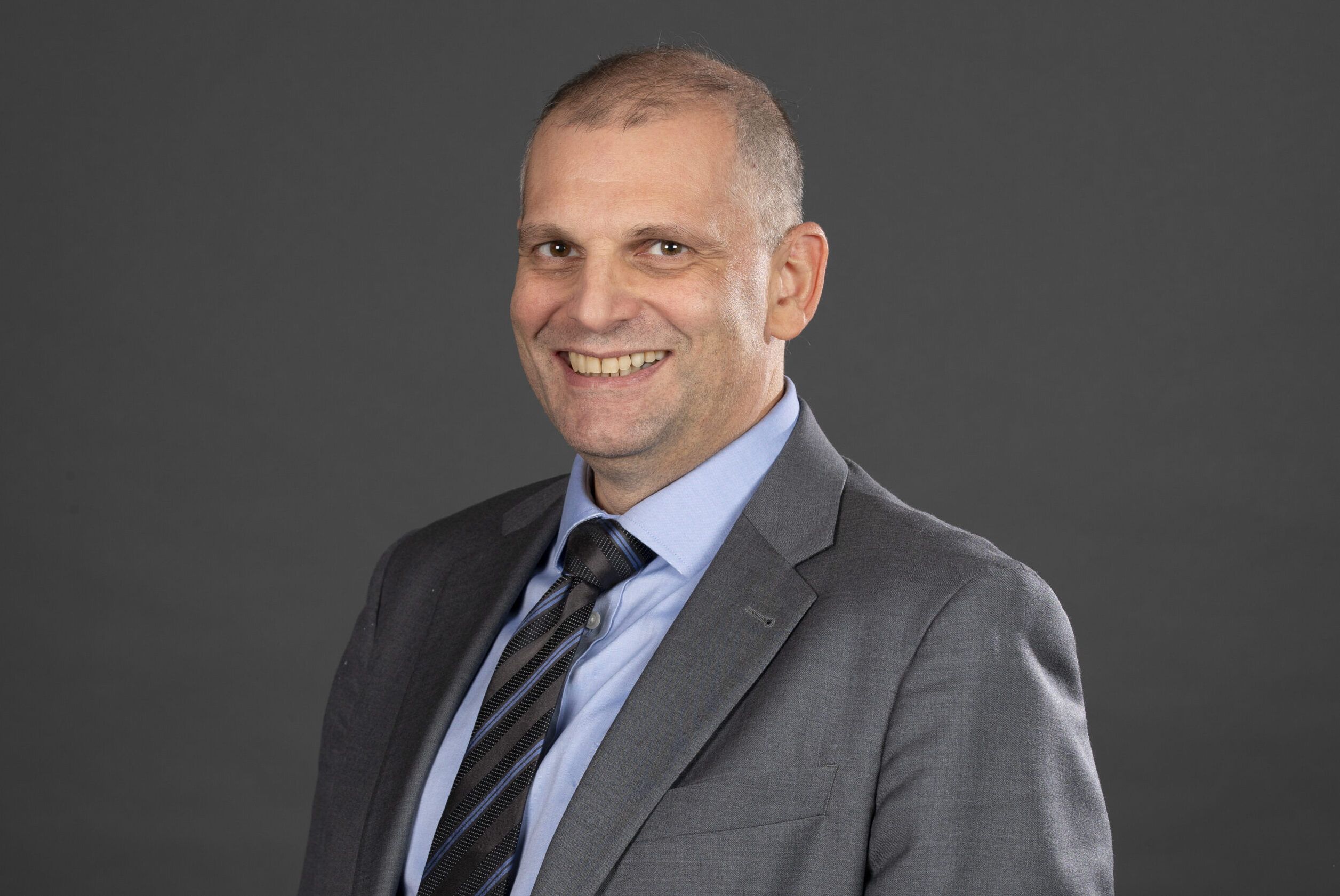 A man in a suit smiles against a dark gray background.