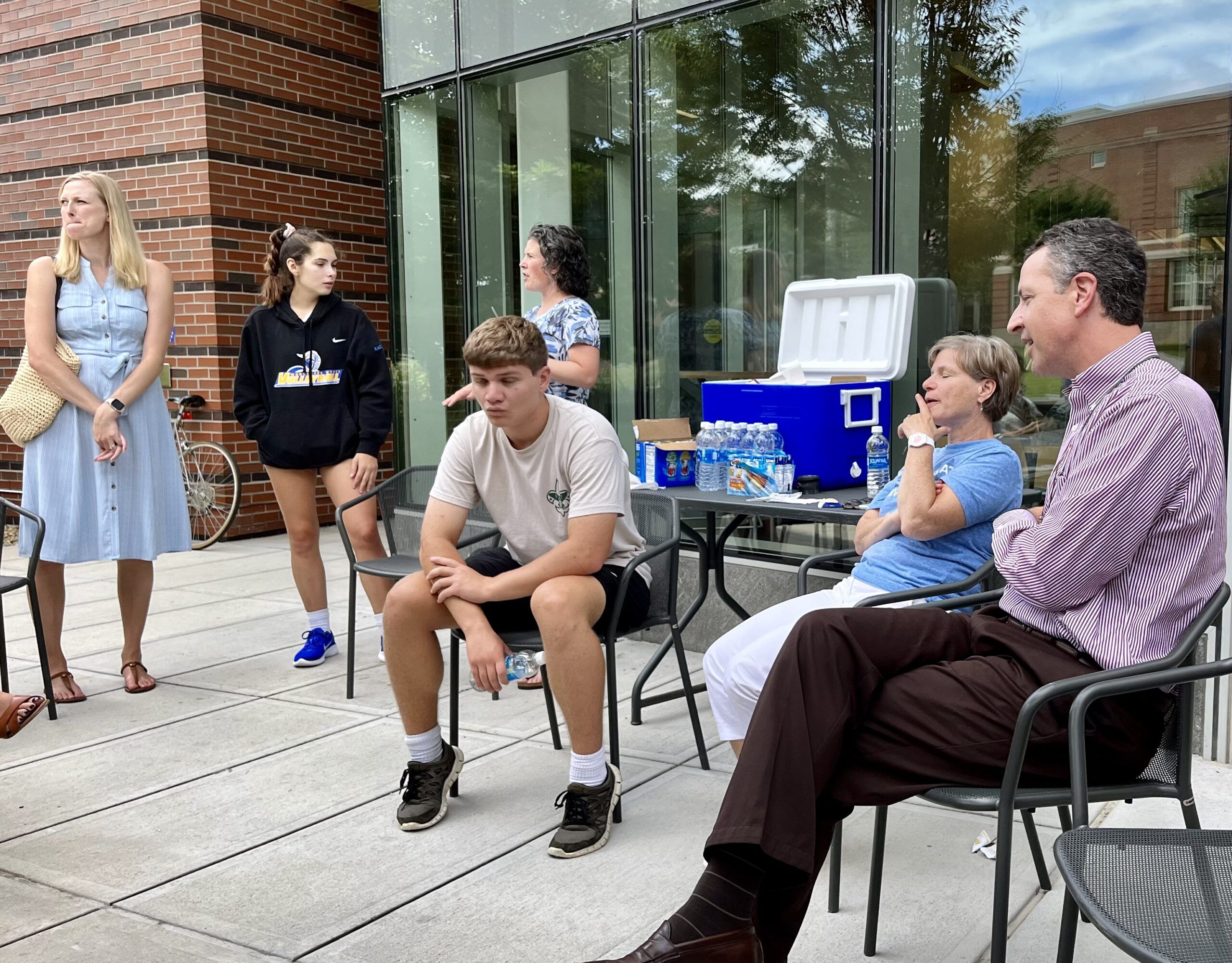 men and woman sitting and standing outside, talking, windows as the background