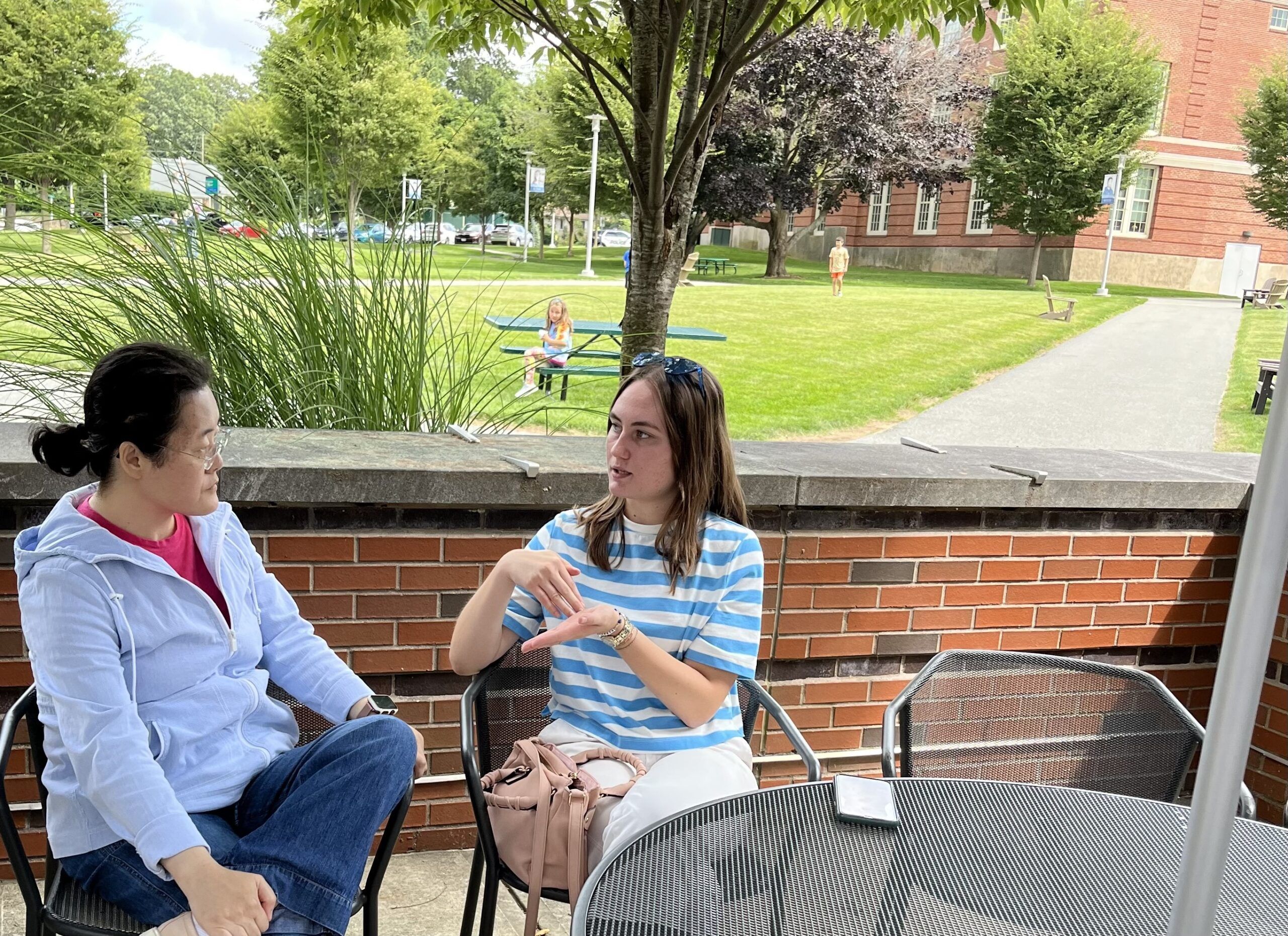 two woman talking and sitting outside. green grass and trees, and brick building as the background