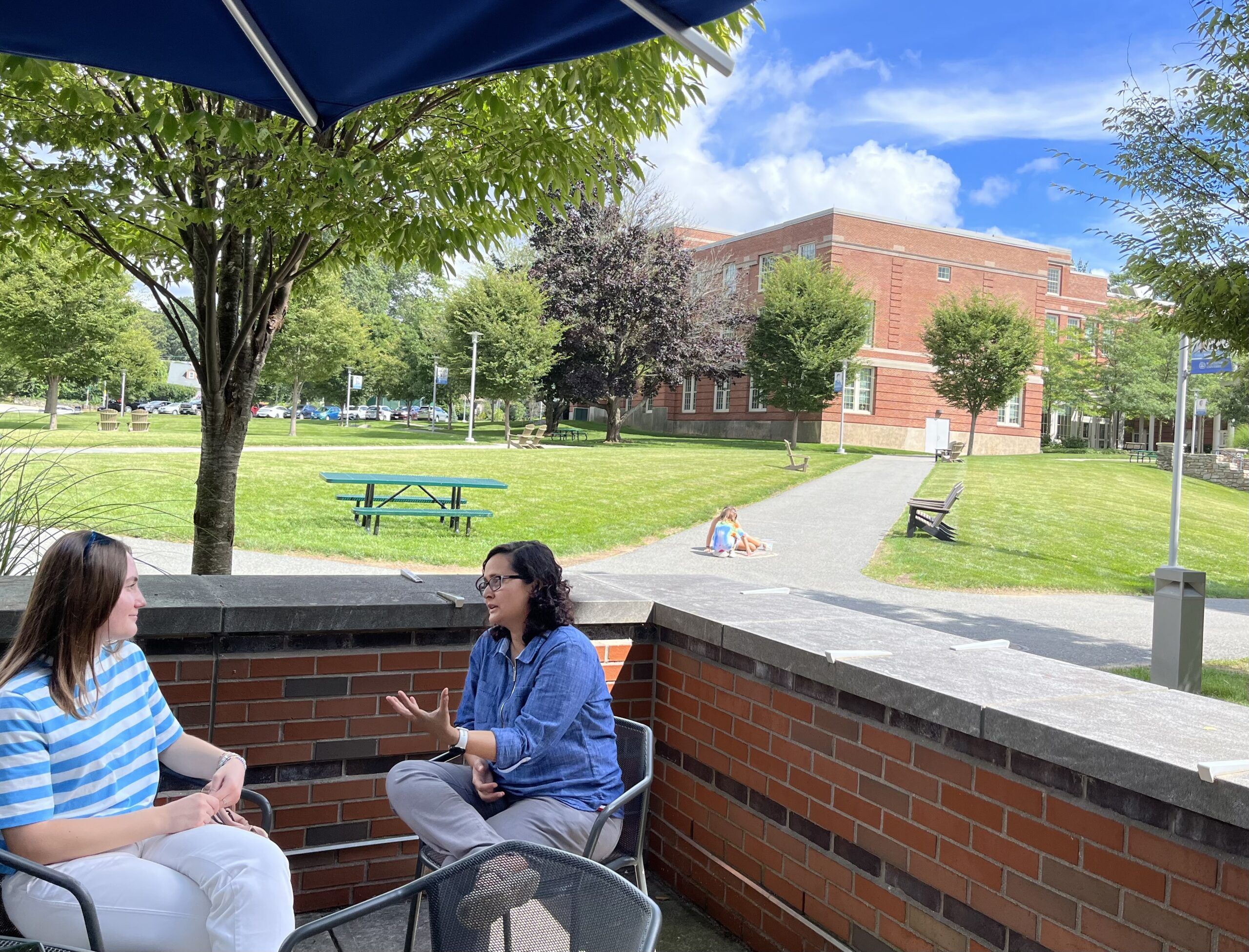 two woman in blue shirts talking, sitting outside, under an umbrella. Blue skies, green grass and trees, and a brick building as the background