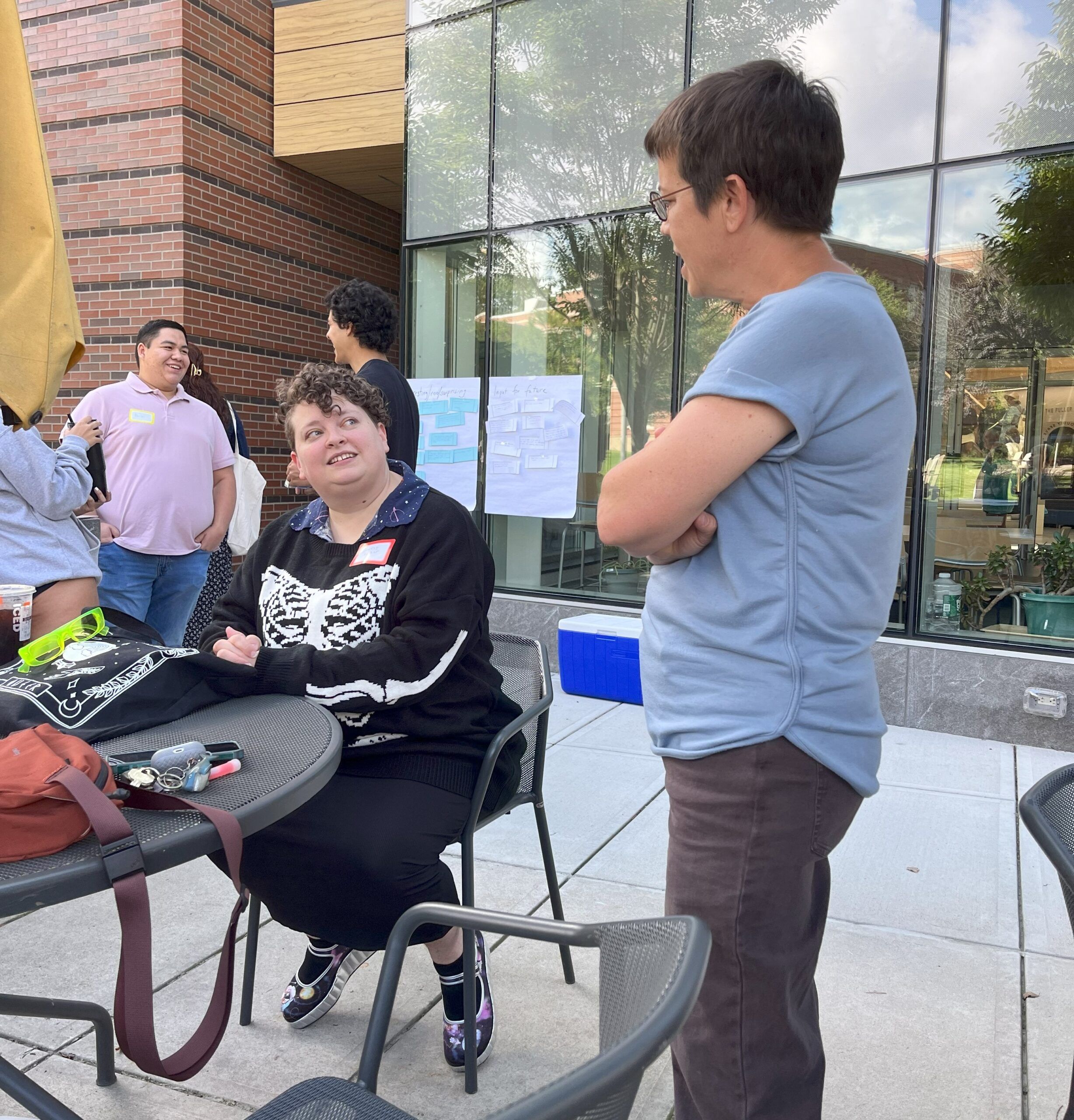 two people talking, one sitting at a table, one standing. windows in the background. three people in the background standing and talking