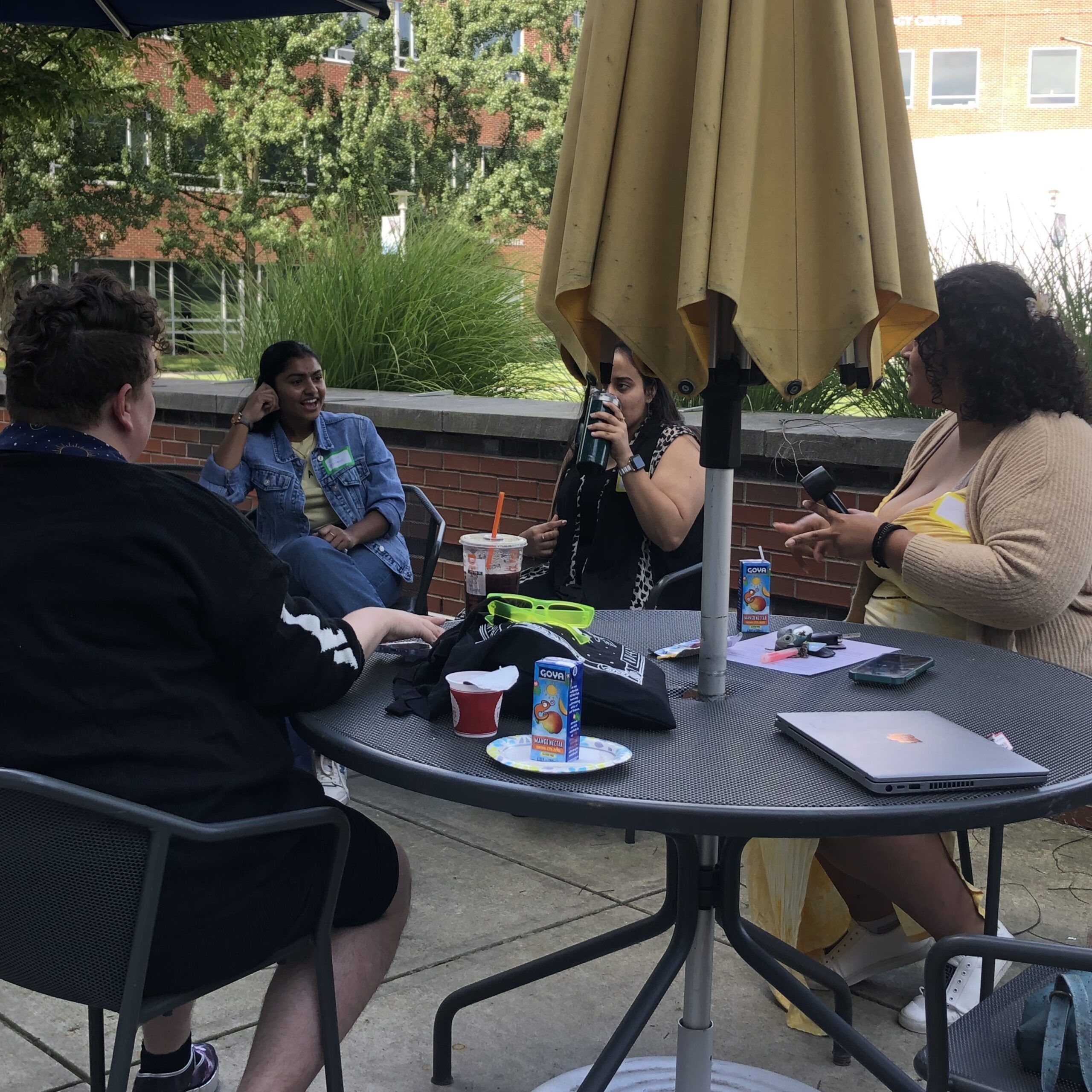 a group of people talking and sitting outside at a black patio table with a light tan table umbrella. Laptops and juice on the table