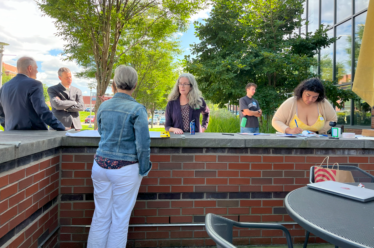 a group of people talking outside, a brick wall and green trees in the background