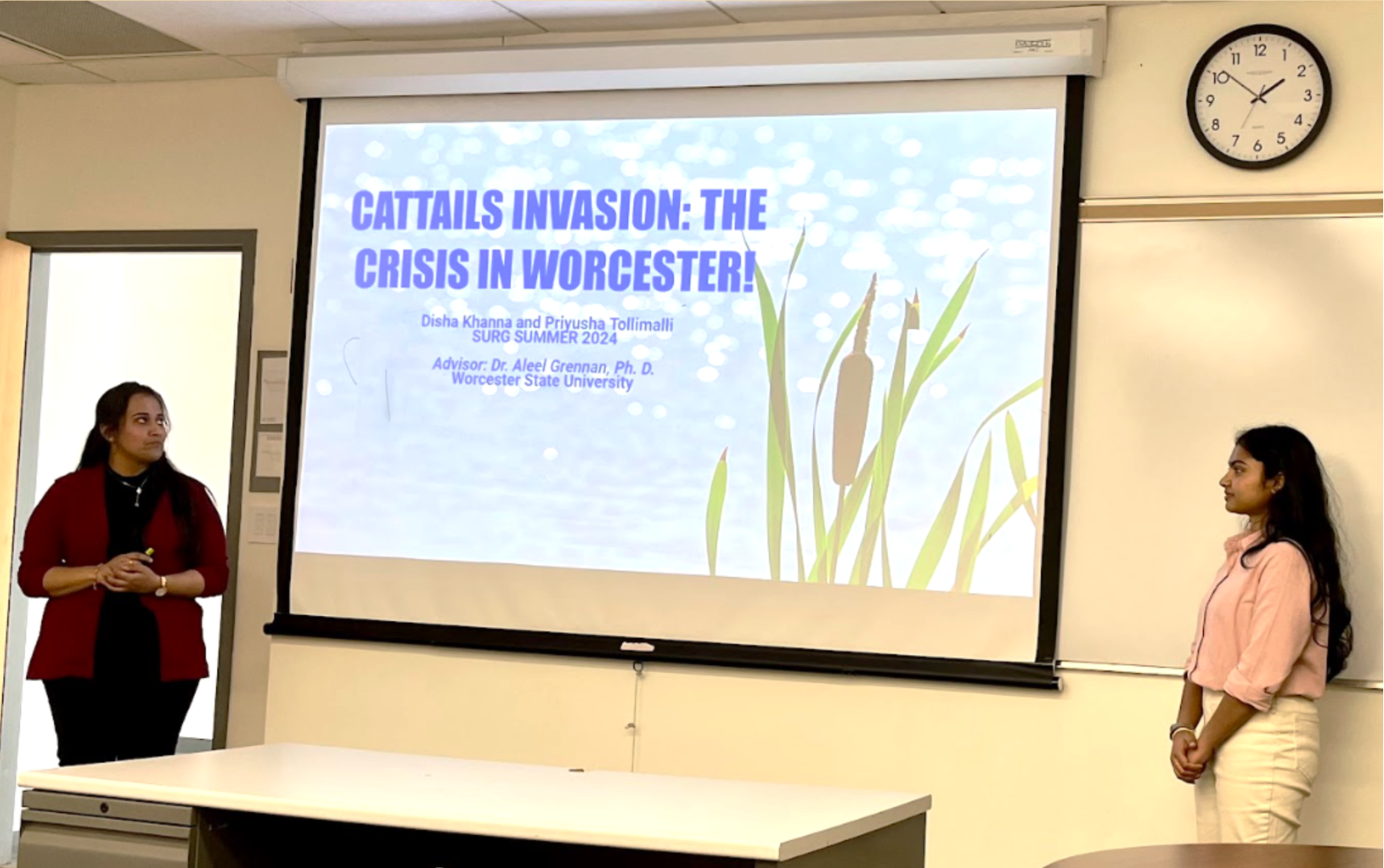 2 women, Priyusha Tollimalli and Disha Khanna in a classroom setting, standing with a projected screen of a presentation between them. Text on the presentation reads "Cattails Invasion: The Crisis in Worcester"